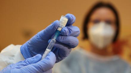 Impfstoff wird in einer Spritze aufgezogen, Frau mit Maske im Hintergrund beobachtet.