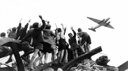West-Berliner Jungen, die auf einem Trümmerberg stehen, begrüßen winkend ein US-amerikanisches Transportflugzeug.