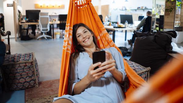 Happy businesswoman holding smart phone lying in hammock in office