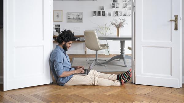 Ein Mann sitzt zu Hause auf dem Boden, mit einem Laptop in seinem Schoß.