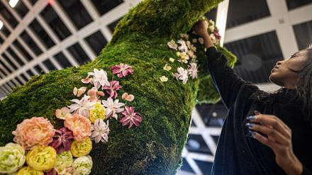 Eine Frau befestigt in der Blumenhalle auf der Internationalen Grünen Woche verschiedene Blumen an der Moos-Skulptur eines Bären.  