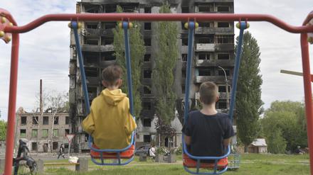 Zwei Kinder sitzen auf einer Schaukel und blicken auf ein durch russische Militärangriffe ausgebranntes mehrstöckiges Wohngebäude.