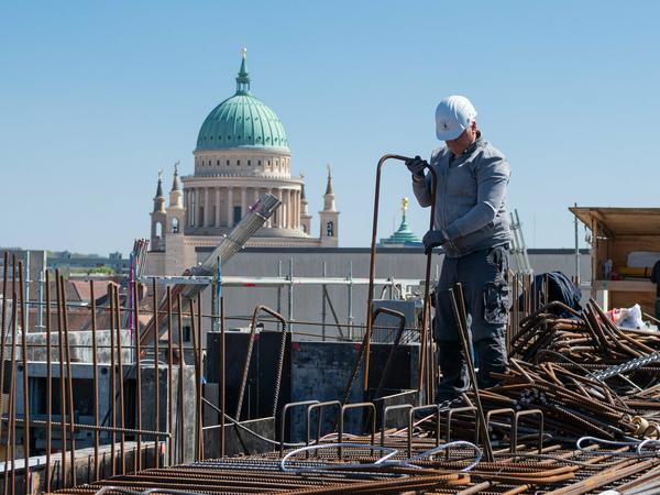 Auf dem Dach des Sockelbaus arbeitet ein Stahl- und Betonbauer an der Bewehrung für das Gießen der Betondecke. Im Hintergrund ist die Kuppel der Kirche St. Nikolai zu sehen. 