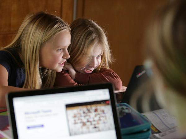 Zwei Mädchen lernen lächelnd vor ihrem Laptop, ihre Mutter arbeitet gegenüber am Computer.